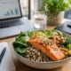 Brainfood-Konzept, gesunde eiweißreiche Mahlzeit auf dem Schreibtisch, Laptop und Notizbuch im Hintergrund, Lachs, Quinoa, grünes Gemüse, Nüsse, weiches natürliches Licht, produktive Stimmung, moderner Büro-Lifestyle
