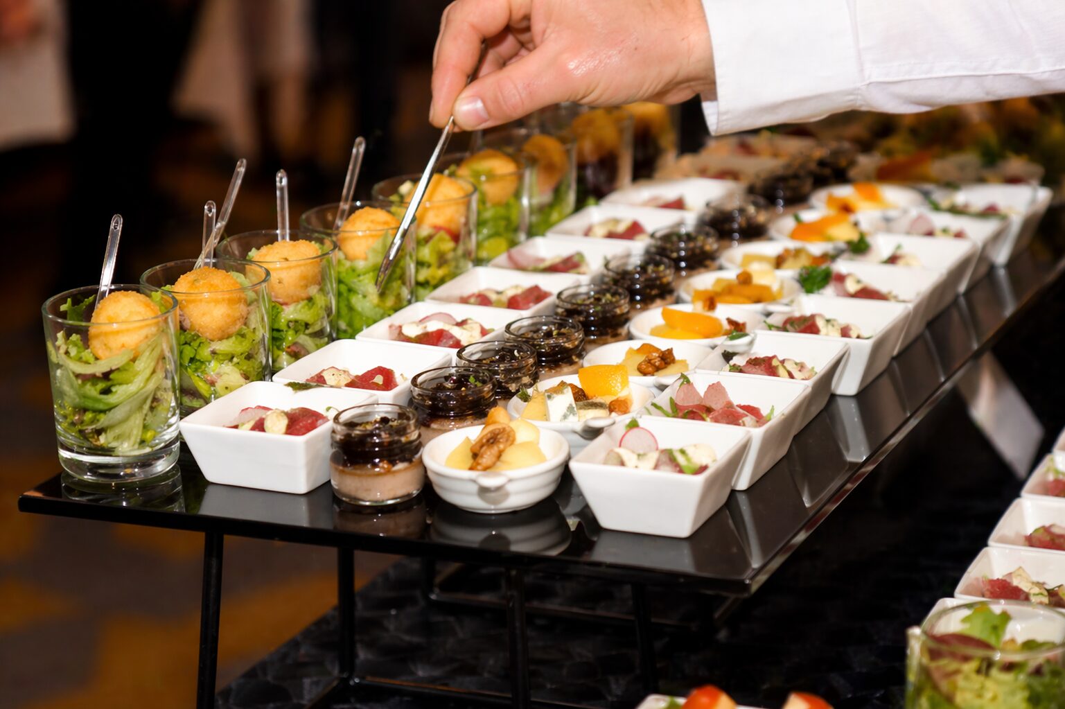 Detailaufnahme eines eleganten Catering-Buffets mit kunstvoll angerichteten Fingerfood-Portionen in Gläsern und Schälchen, während eine Person mit einer Pinzette ein Häppchen auswählt.